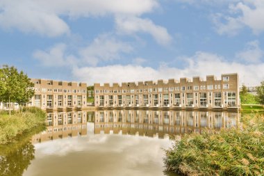 Su kenarında mavi gökyüzü ve beyaz bulutlarla çevrili bir apartman dairesi havuzun karşısından çekiliyor.