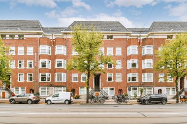 Pencerelerinde beyaz süsleme olan tuğla bir binanın önüne park etmiş birkaç araba ve caddeyi çevreleyen yeşil ağaçlar.