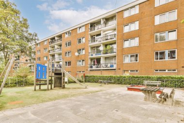 Önünde oyun alanı olan tuğladan bir apartman ve binanın diğer tarafında ağaçlar olan bir açık hava oyun alanı var.