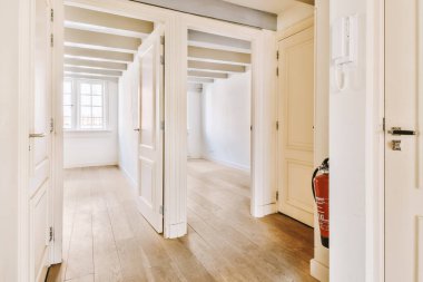 Beyaz duvarları ve sağ tarafında ahşap döşemeleri olan boş bir oda. Kapıda kırmızı bir yangın çıkışı var.