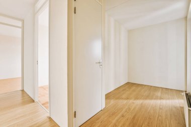 Beyaz duvarları ve sağında ahşap döşemeleri olan boş bir oda. Başka bir odaya açılan bir kapı var.