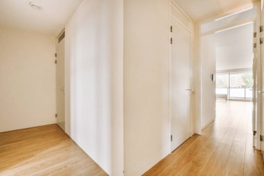 Beyaz duvarları ve sağında ahşap döşemeleri olan boş bir oda. Başka bir odaya açılan bir kapı var.