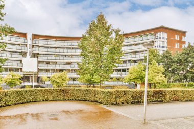 Bulutlu bir günde büyük, çok katlı bir binanın önünde ağaçların ve çalıların olduğu bir alan.