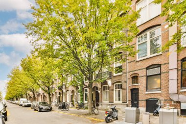 Caddenin her iki tarafında da park etmiş arabalar ve ağaçlarla dolu bir şehir caddesi var. Arka planda mavi bir gökyüzü var.