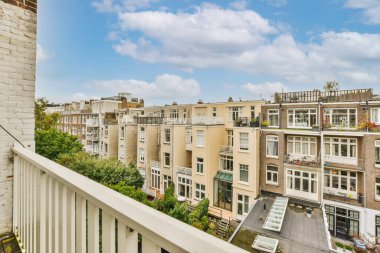 Arka planda bir sürü ev ve üzerinde mavi gökyüzü olan bir balkon bir apartman penceresinden görüldüğü gibi.