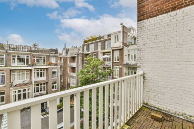 Parlak güneşli bir günde arka planda beyaz korkuluklu ve tuğlalı binaların olduğu bir balkon.