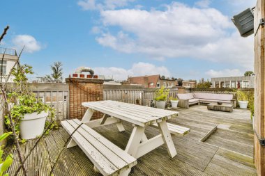 Fotoğrafta ahşap güverte ve beyaz piknik masaları olan bir açık hava alanı çatıdan arka bahçeye çekiliyor.