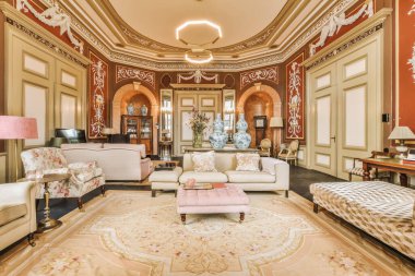 Oturma odası, koltuklar, sandalyeler ve halılar süslü bir duvarın önünde.