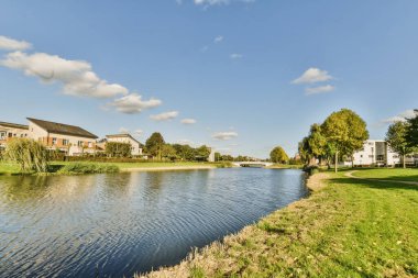 Arka planda evleri ve üzerinde mavi gökyüzü olan bir göl, nehrin karşısından görülebildiği gibi beyaz tüylü bulutlardır.