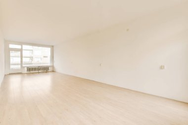 Ahşap döşemeleri ve beyaz duvarları olan boş bir oturma odası, sağdaki duvarda bir pencere var.