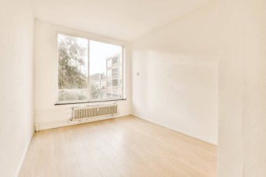 Beyaz duvarları ve sağında ahşap döşemeleri olan boş bir oda, solunda büyük bir pencere var.