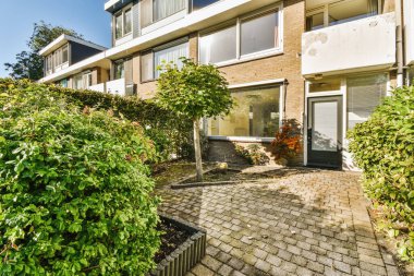 Önünde ağaçlar ve çalılar olan bir apartmanın önünde güneşli mavi bir gökyüzünde