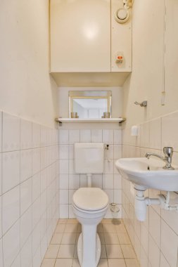 Duvarlarında ve zemininde beyaz fayanslar olan küçük bir banyo. Köşedeki tuvaletin üstünde bir ayna var.