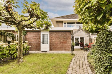 Ön bahçesinde tuğla duvarlar ve yeşil çimenler olan bir ev, yanında büyüyen bir ağaç var.