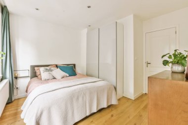 Beyaz duvarları ve ahşap zemini olan bir yatak odası büyük pencerelerden gelen doğal ışıkla aydınlanır.