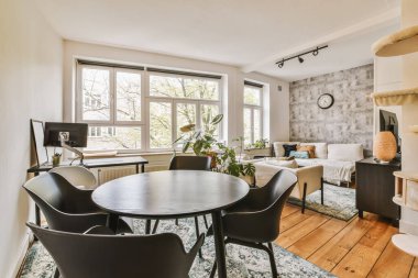 Güneşli bir günde pencerenin önünde sandalyeli ve yuvarlak masalı bir oturma odası bahçeye bakıyor.