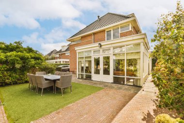 Güneşli bir günde yemyeşil ağaçlarla çevrili, arka bahçesinde bir avlusu ve yemek alanı olan bir ev.