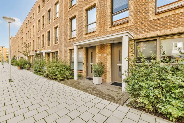 Her iki tarafında bitkiler ve çalılar olan tuğla bir bina. Kapının yanında sokak lambası direği var.