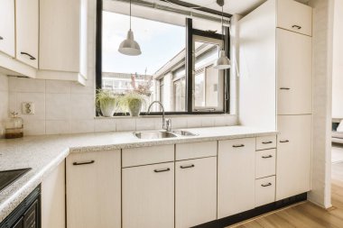 Pencerenin etrafında beyaz dolaplar ve siyah süslemeler olan mutfak alanı lavabonun önündedir.