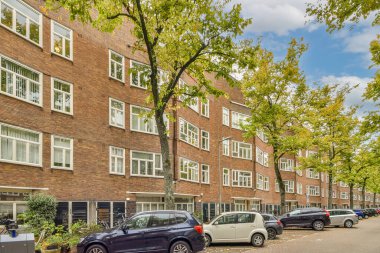 Bazı arabalar sokağın kenarına park edilmiş. Tuğla bir binanın önünde. Bir sürü pencere ve balya var.