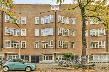 Tuğladan bir apartmanın önüne park edilmiş yeşil bir araba. Üst katında bir sürü pencere ve balkon var.