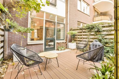 İki sandalyeli bir teras ve güvertede bir masa, ağaçlarla çevrili tuğla bir binanın önünde.