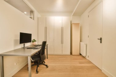 Masasında bilgisayar olan boş bir oda ve solunda duvarda iki beyaz dolap.