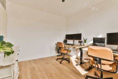 Duvarında iki masa ve üç bilgisayar ekranı olan bir ofis. Birinin içinde yeşil bir bitki var.