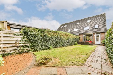 Evin her iki tarafında da tuğla kaldırımlar ve yeşil bitkilerle dolu bir arka bahçesi var. Arka planda mavi bir gökyüzü var.