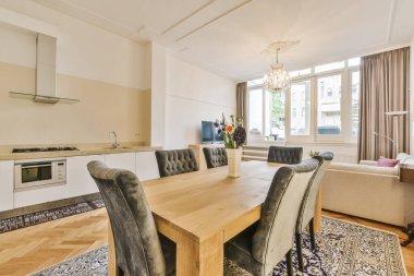 Ahşap döşemesi, beyaz duvarları, bej perdeleri, ahşap masası ve sandalyeleri olan bir dairede oturma odası ve yemek odası.