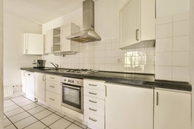 Bu fotoğrafta tezgahların üzerinde beyaz dolaplar ve siyah tezgahlar olan bir mutfak içeriden çekilmiş.