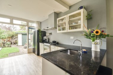 Siyah tezgahlı bir mutfak ve arkasındaki beyaz dolaplar bahçeye açılan açık cam bir kapı.