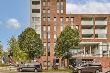 Uzun tuğlalı bir binanın önüne park etmiş iki araba ve caddelerde kuyruk oluşturan ağaçlar.