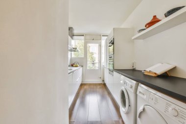 Bölgede ahşap döşeme ve beyaz çamaşır makinesi olan bir çamaşır odası pencereden gelen doğal ışıkla aydınlanır.