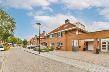 Yan tarafında arabalar olan boş bir cadde ve sokağın ortasında ağaçlarla çevrili tuğla binalar.