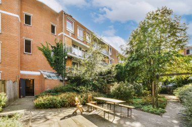 Güneşli bir mavi gökyüzü gününde tuğla bir apartmanın önünde masaların, sandalyelerin ve ağaçların olduğu açık bir alan.