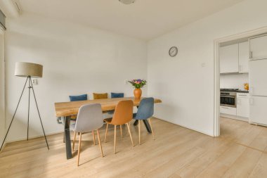 Beyaz duvarlı bir odada yemek masası ve sandalyeler, ahşap döşemeler ve sağ tarafında açık renkli mobilyalar.