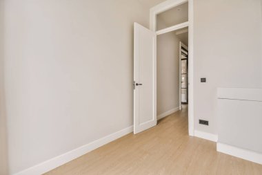 Odanın ortasında beyaz duvarları ve ahşap döşemeleri olan boş bir oda, başka bir odaya açılan bir kapı var.