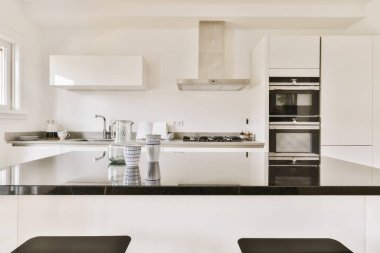 Odanın ortasında siyah tezgahları ve tabureleri olan beyaz bir mutfak yemek alanına bakıyor.