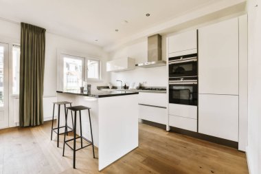 Adanın önünde beyaz dolapları ve siyah tezgahları olan modern bir mutfak pencerenin yanındadır.