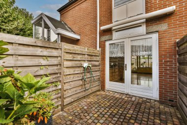 Tuğla duvarı ve yeşil bitkilerle çevrili açık bir avlu alanına açılan beyaz kapısı olan bir evin dışında.