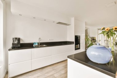 Odanın ortasında beyaz dolapları ve siyah tezgahları olan modern bir mutfak, çiçeklerle dolu büyük mavi bir vazo.