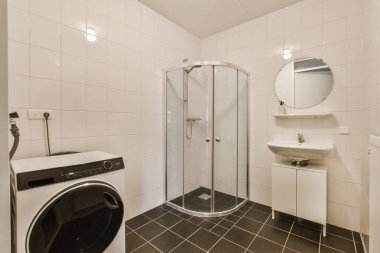 Banyonun köşesinde çamaşır makinesi ve kurutma makinesi olan küçük bir banyo küvetin yanında beyaz fayanslar var.