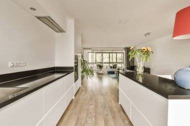 Odanın ortasında beyaz dolapları ve siyah tezgahları olan modern bir mutfak turuncu bir kolye ışığı var.