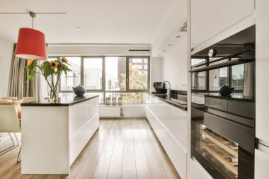 Odanın ortasında ahşap döşemeli ve beyaz dolaplı modern bir mutfak adanın üzerinde asılı kırmızı bir lamba var.