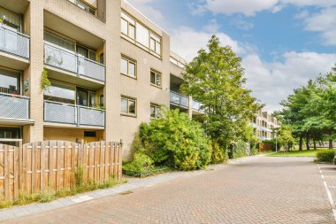 Tuğla bir binanın önünde boş bir cadde. Kenarları baldüğümlü ve kaldırımları ağaçlarla kaplı.