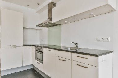 Bu fotoğrafta tezgahların üzerinde beyaz dolaplar ve siyah tezgahlar olan bir mutfak içeriden çekilmiş.