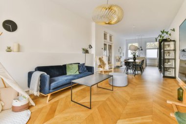 Odanın ortasında ahşap döşemeli ve beyaz duvarlı bir oturma odası mavi kadife bir kanepe.