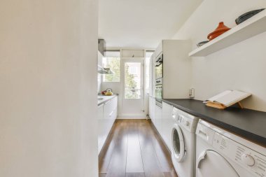 Bölgede ahşap döşeme ve beyaz çamaşır makinesi olan bir çamaşır odası pencereden gelen doğal ışıkla aydınlanır.