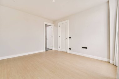 Beyaz duvarları ve sağında ahşap döşemeleri olan boş bir oda. Başka bir odaya açılan bir kapı var.